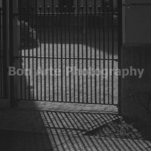 A high contrast black and white fine art photograph of a heavy black gate casting a stark linear geometric shadow pattern across a cobblestone pavement for music album cover photography by Bon Arte Photography.