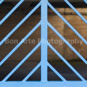 A sharp close up of a bright blue painted metal security gate with repeating diagonal grille lines forming a chevron pattern against a warm blurred concrete background for electronic music album cover photography by Bon Arte Photography.
