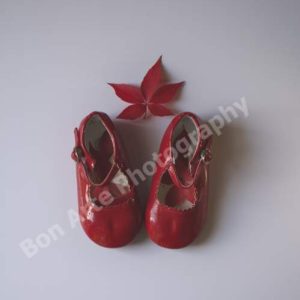 An original concept print featuring a striking red shoes photo, showcasing a pair of vintage patent leather children's shoes with a single crimson vine leaf centered above them on an off-white background.