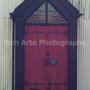 A weathered gothic red door with a dark triangular wooden frame set against a corrugated white wall for thriller book cover design by Bon Arte Photography.