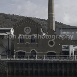 A muted, desaturated photograph of an old brick industrial warehouse building with a tall brick factory chimney stack by a riverfront, designed with negative space for thriller novel typography.