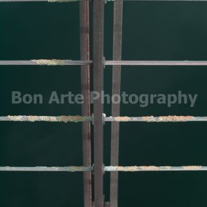 A minimalist vertical geometric composition of a metal dock post and wire cable railing covered in green marine algae crust over dark coastal sea water, ideal for thriller title layouts.