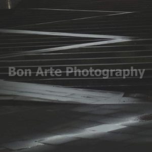A dark, desaturated square photograph of outdoor concrete steps featuring a prominent, geometric zigzag ramp cutting through the stairs with dramatic light reflections