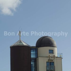 A minimalist architectural composition showing a dark astronomy observatory dome next to a tower topped with a classical female statue holding a hoop against a clear blue sky for ambient electronic music album cover photography by Bon Arte Photography.