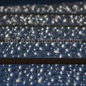 A macro close up shot of pristine rain drops beaded on horizontal dark wooden slats with shifting bokeh light reflections for ambient music album cover photography by Bon Arte Photography.