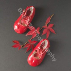 A pair of glossy red children shoes arranged as if taking footsteps next to a branch of vibrant red autumn leaves on a flat dark grey background for thriller novel book cover photography by Bon Arte Photography.