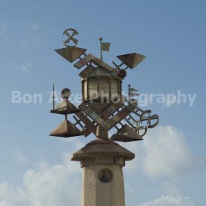 A complex steampunk kinetic weather vane sculpture made of copper metal pyramids gears and windmill vanes atop a stone pillar against a bright blue sky for industrial electronic music album cover photography by Bon Arte Photography.